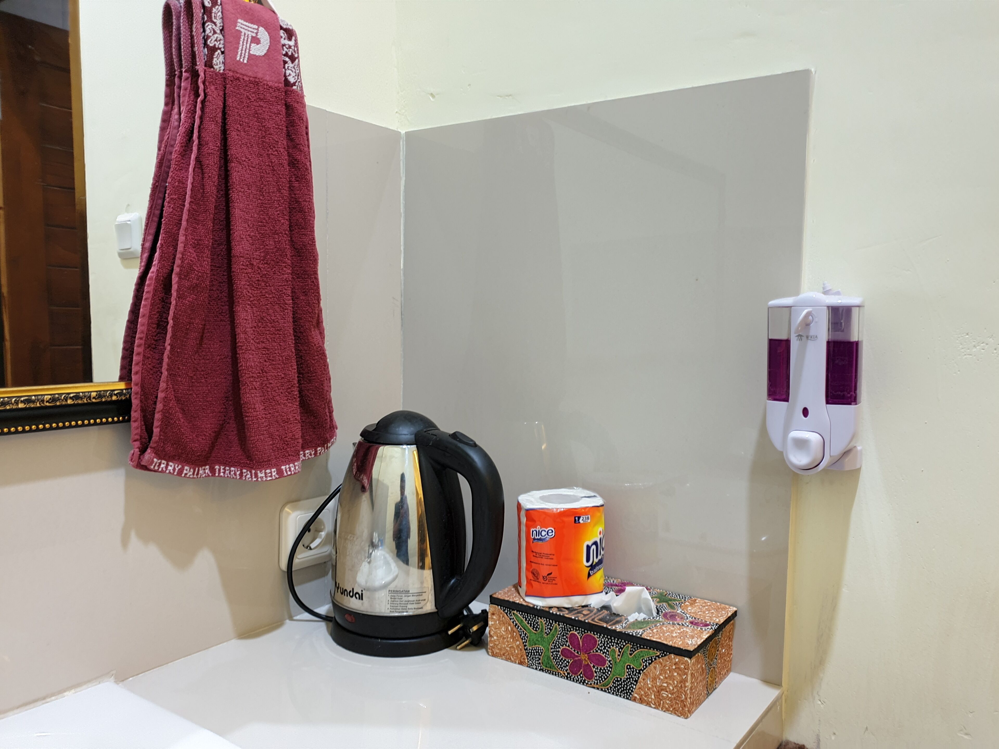 Ein Badezimmer mit einem weißen Waschbecken, einem roten Handtuch an der Wand, einem Wasserkocher, einer Toilettenpapierpackung und einer Seifenspender.