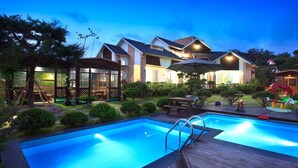 A swimming pool with a wooden deck, surrounded by a well-maintained garden and a house with a sloped roof. - Gyeongju Orsay Pension (Gyeongju)