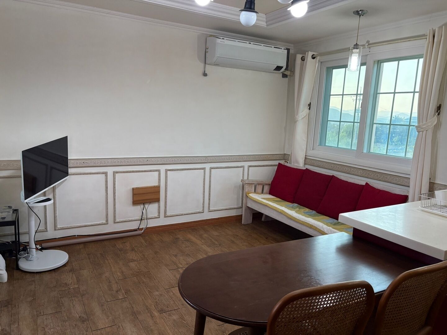 Una habitación con piso de madera, una mesa de comedor, un banco con cojines rojos, un televisor montado en la pared y una ventana con cortinas.