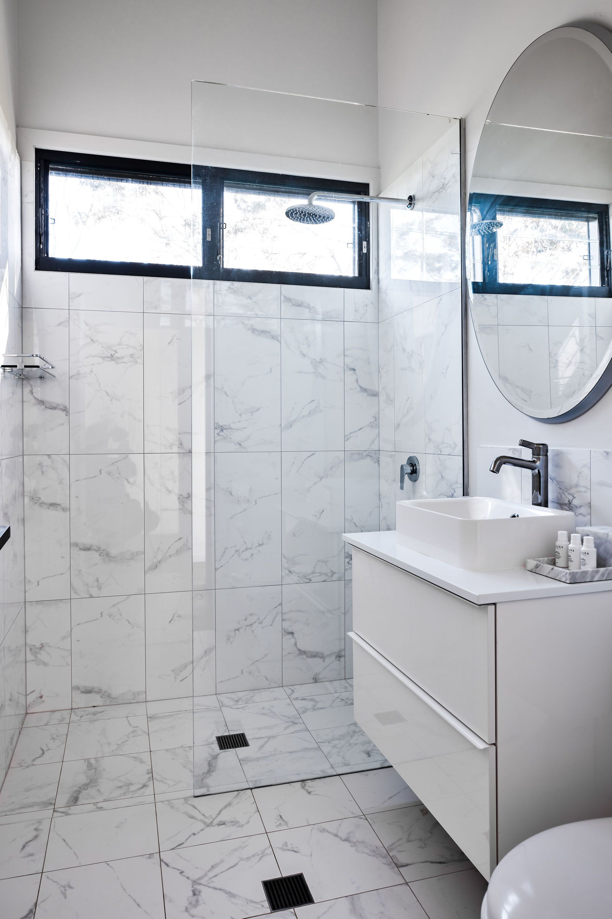 Un baño moderno con piso de mármol blanco, un espejo grande, un lavamanos blanco y una ducha con puertas de vidrio.