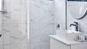 Un baño moderno con piso de mármol blanco, un espejo grande, un lavamanos blanco y una ducha con puertas de vidrio.