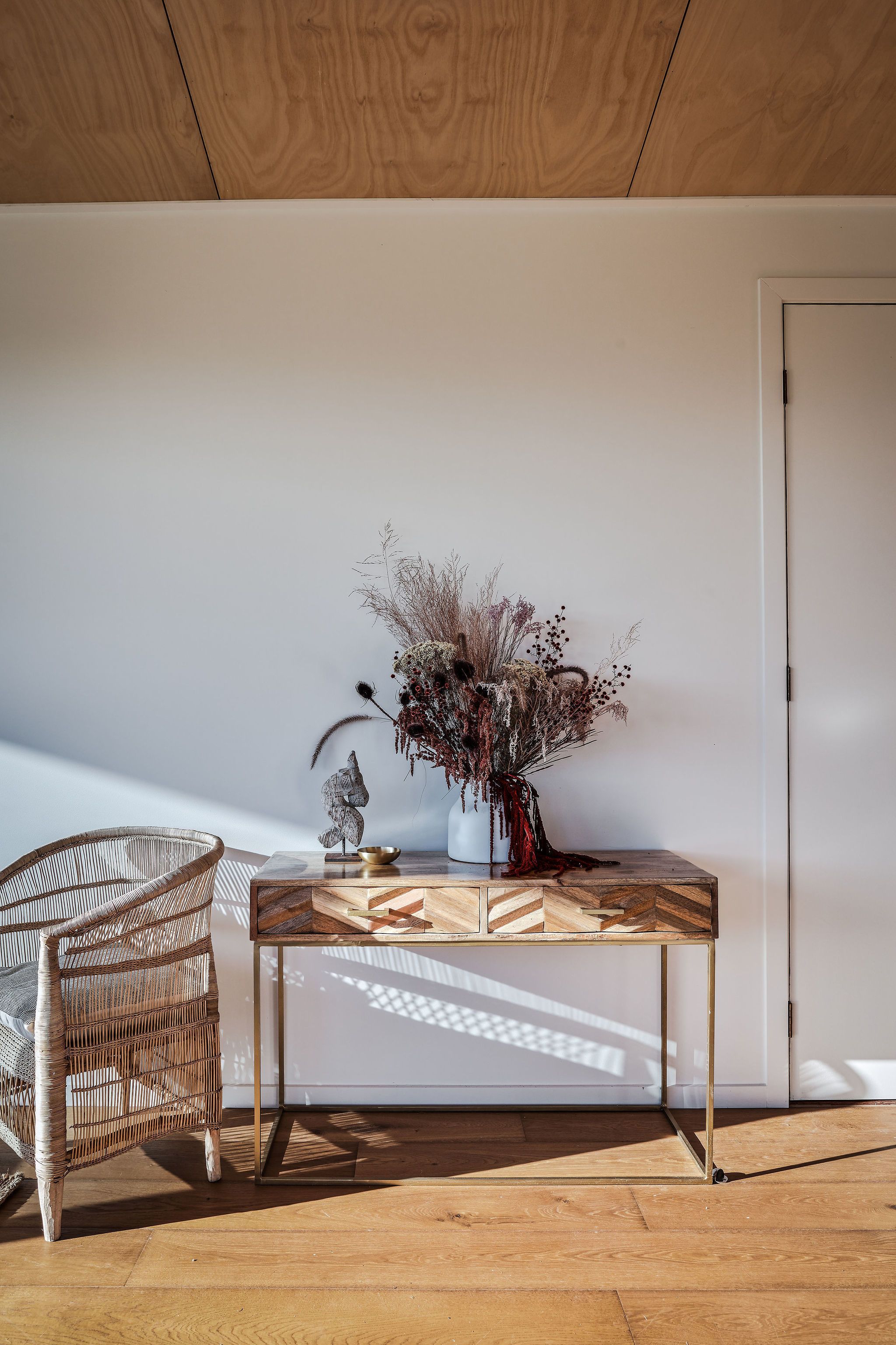 A wooden floor, a wicker chair, a table with a vase of dried flowers, and a white door.
