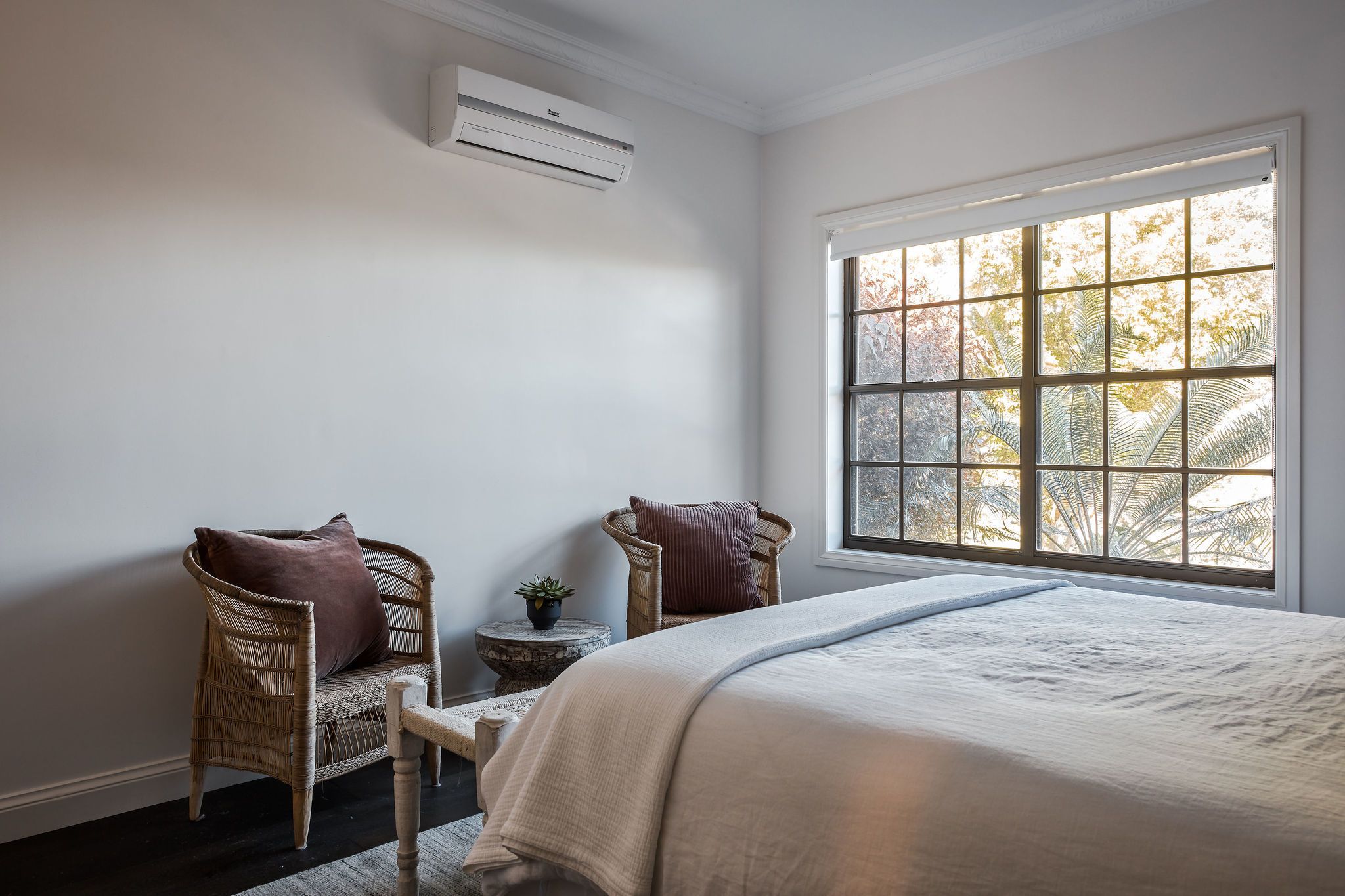 A bedroom with a large window, a bed, two wicker chairs, a small round table, and an air conditioning unit.