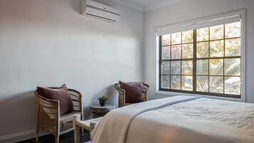 A bedroom with a large window, a bed, two wicker chairs, a small round table, and an air conditioning unit.