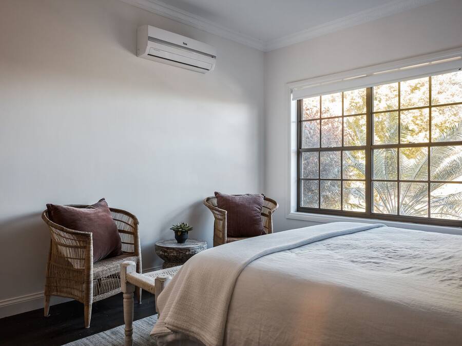A bedroom with a large window, a bed, two wicker chairs, a small round table, and an air conditioning unit.