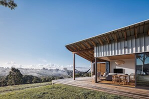 Een modern huis met een houten terras, een hangmat en een televisie in een groot raam.