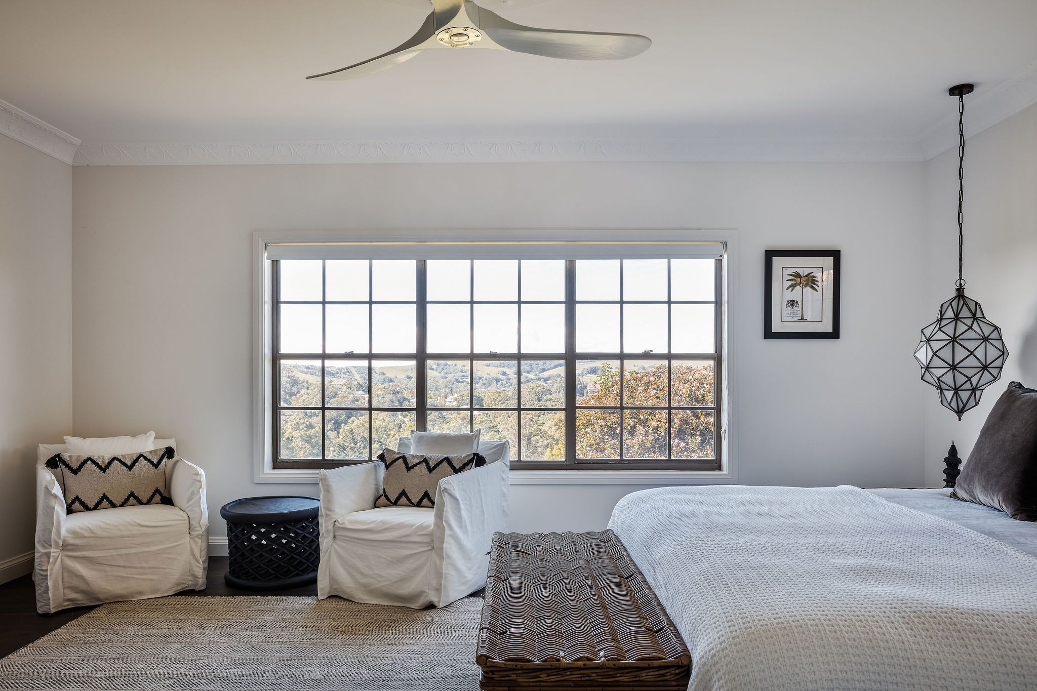 A bedroom with a large window, a bed, two chairs, a small round table, and a hanging light fixture.