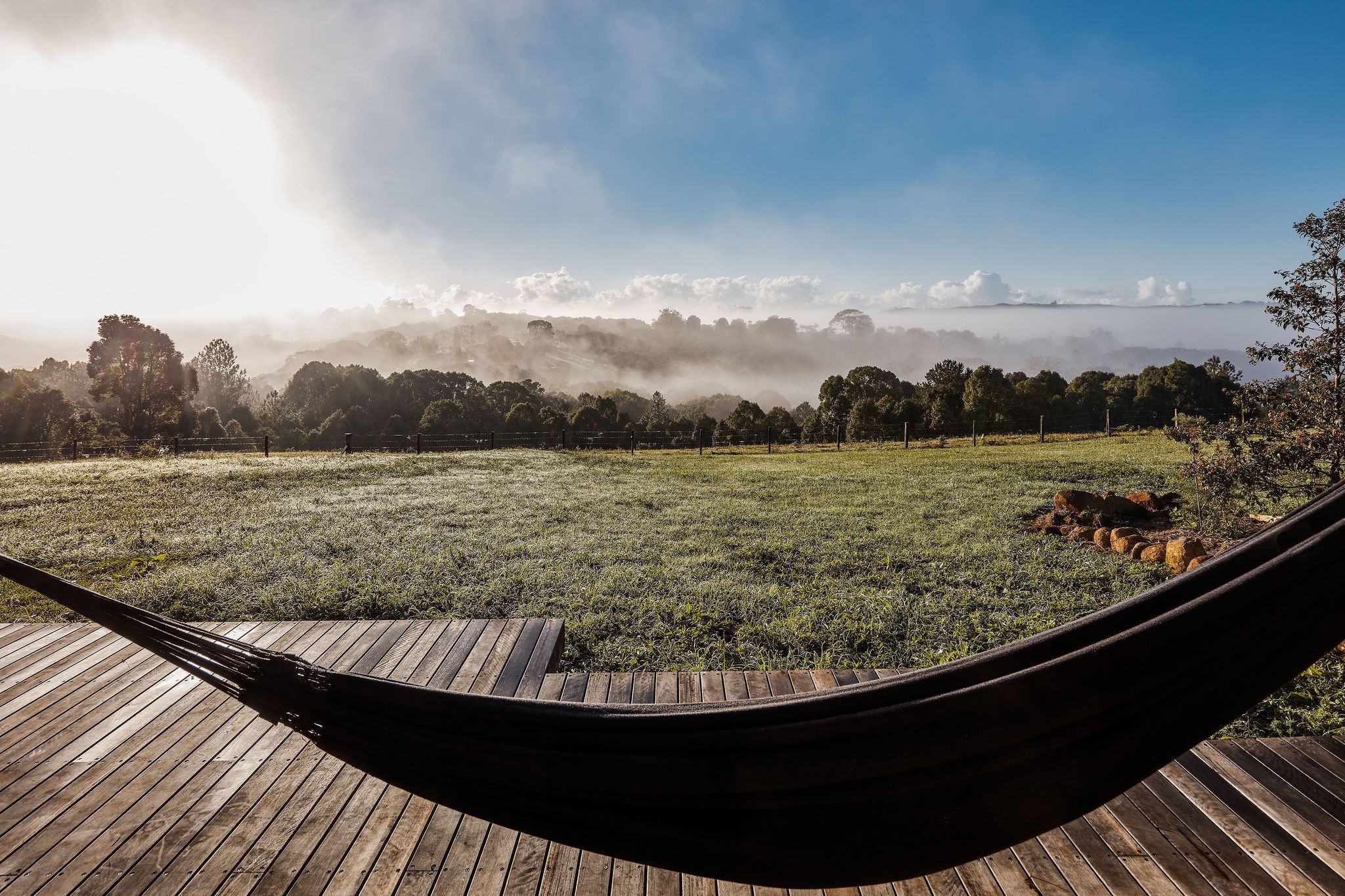 Un hamac sur une terrasse en bois donnant sur un champ verdoyant, avec des arbres et un horizon brumeux.
