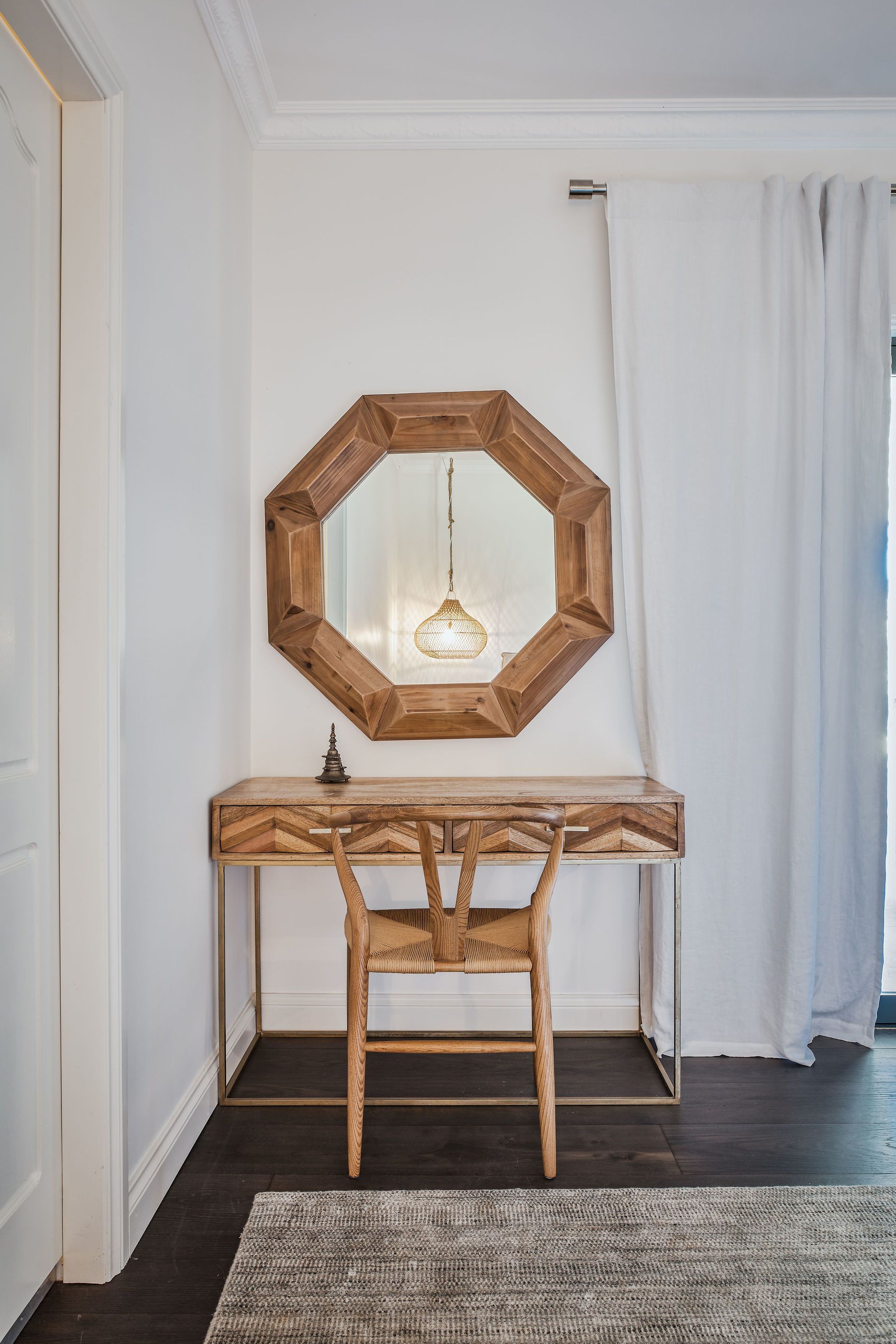 A wooden console table with a geometric mirror above it, a woven chair, and a textured rug on a dark floor.