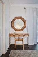 A wooden console table with a geometric mirror above it, a woven chair, and a textured rug on a dark floor.