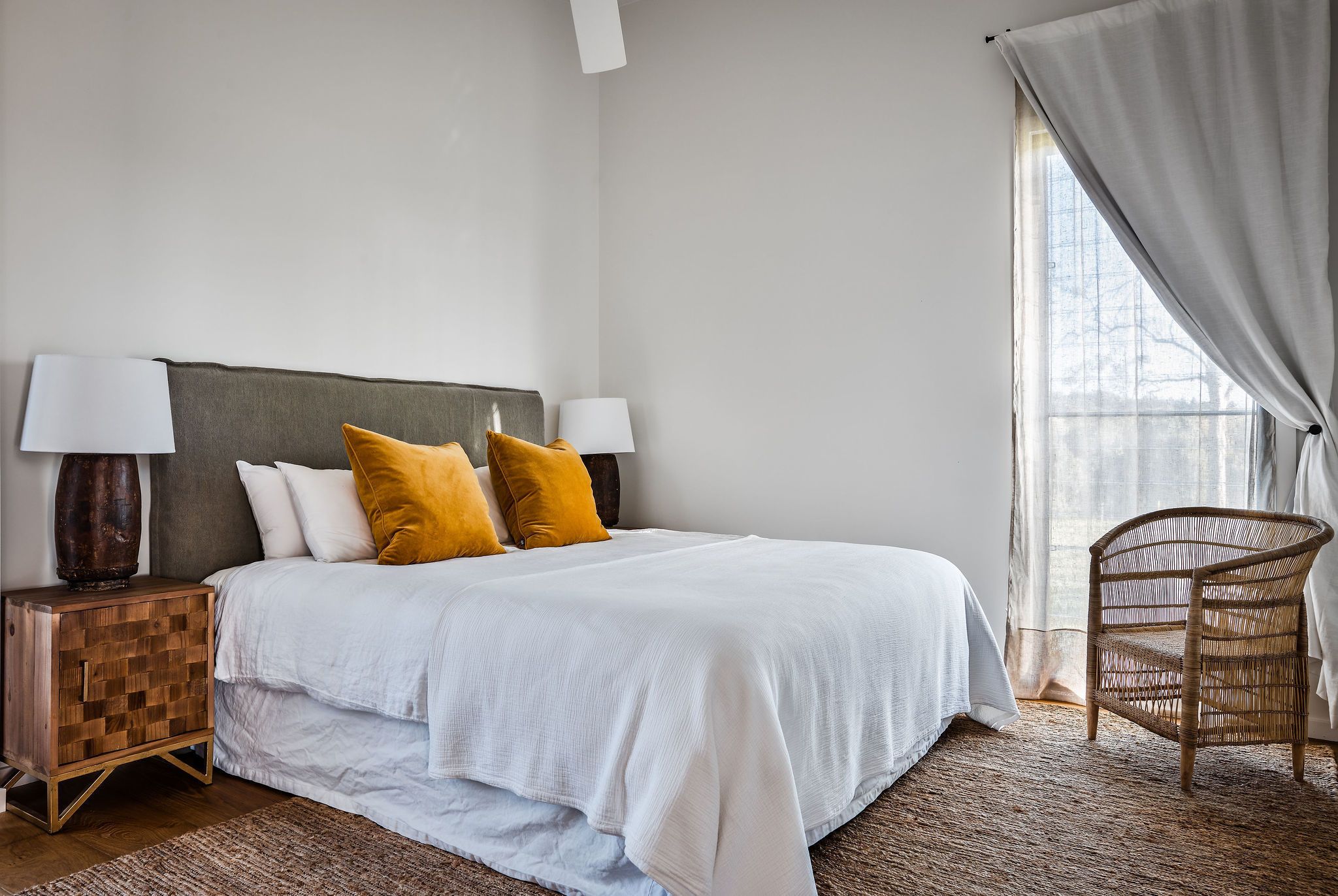 Une chambre à coucher bien rangée, avec un lit, des lampes de chevet, un fauteuil en osier et une fenêtre avec des rideaux.
