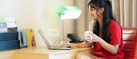 Una persona sentada en un escritorio con una computadora portátil, un teléfono y una taza, en una habitación con una lámpara de escritorio y cortinas.