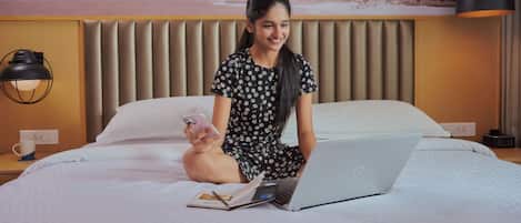 Uma pessoa sentada em uma cama com um laptop, segurando um smartphone e um livro em um quarto de hotel, com uma cama, travesseiros e uma luminária fixada na parede.