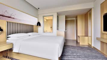 A modern hotel room with a large bed, a bedside table, a TV mounted on the wall, and a bathroom visible through an open door.