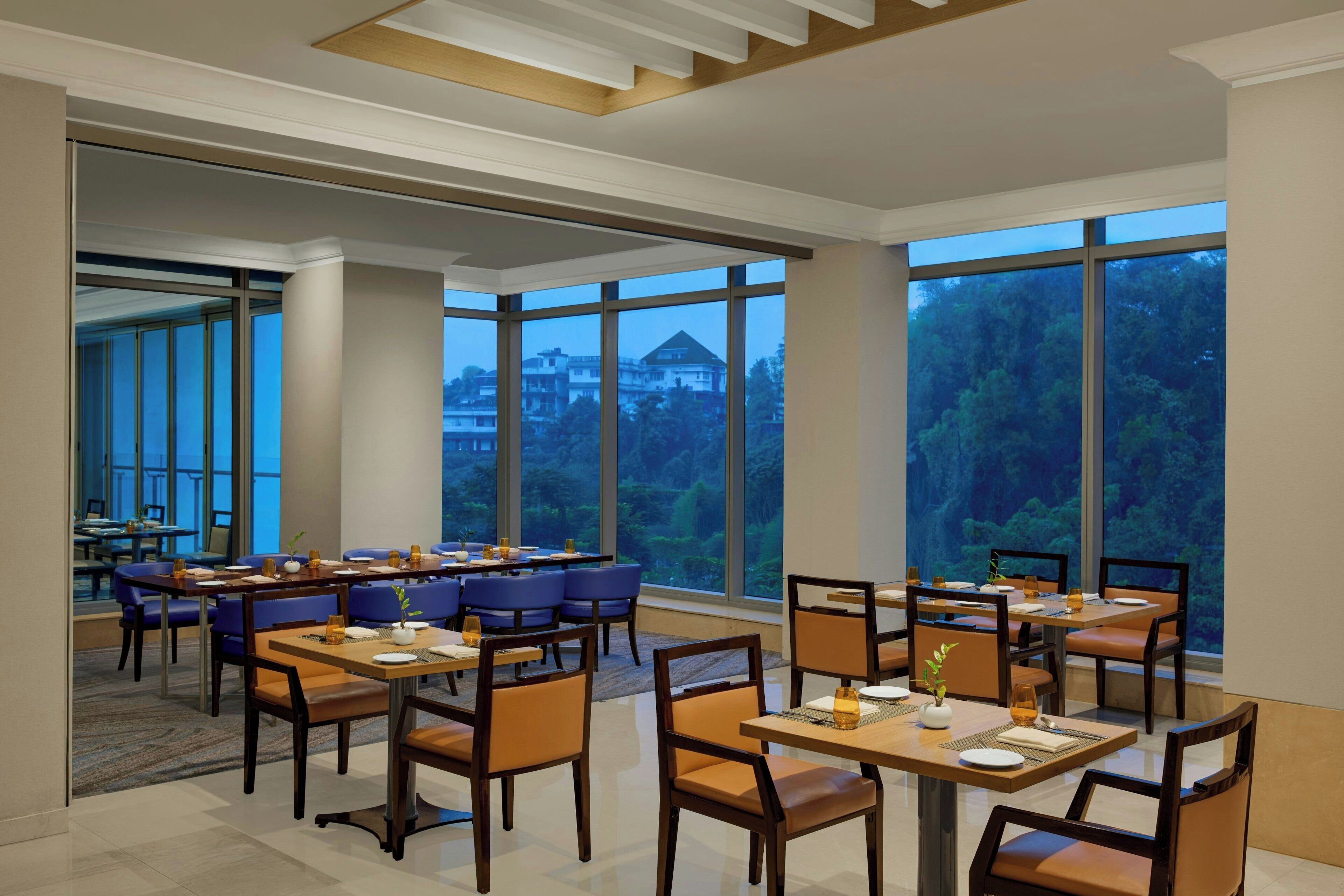 A spacious dining area with multiple tables and chairs, large windows with a view of trees and buildings, and a modern ceiling design.