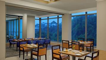 A spacious dining area with multiple tables and chairs, large windows with a view of trees and buildings, and a modern ceiling design.