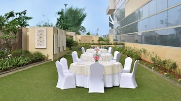An outdoor dining area with round tables and white chairs, surrounded by a well-maintained lawn and a building with large windows.