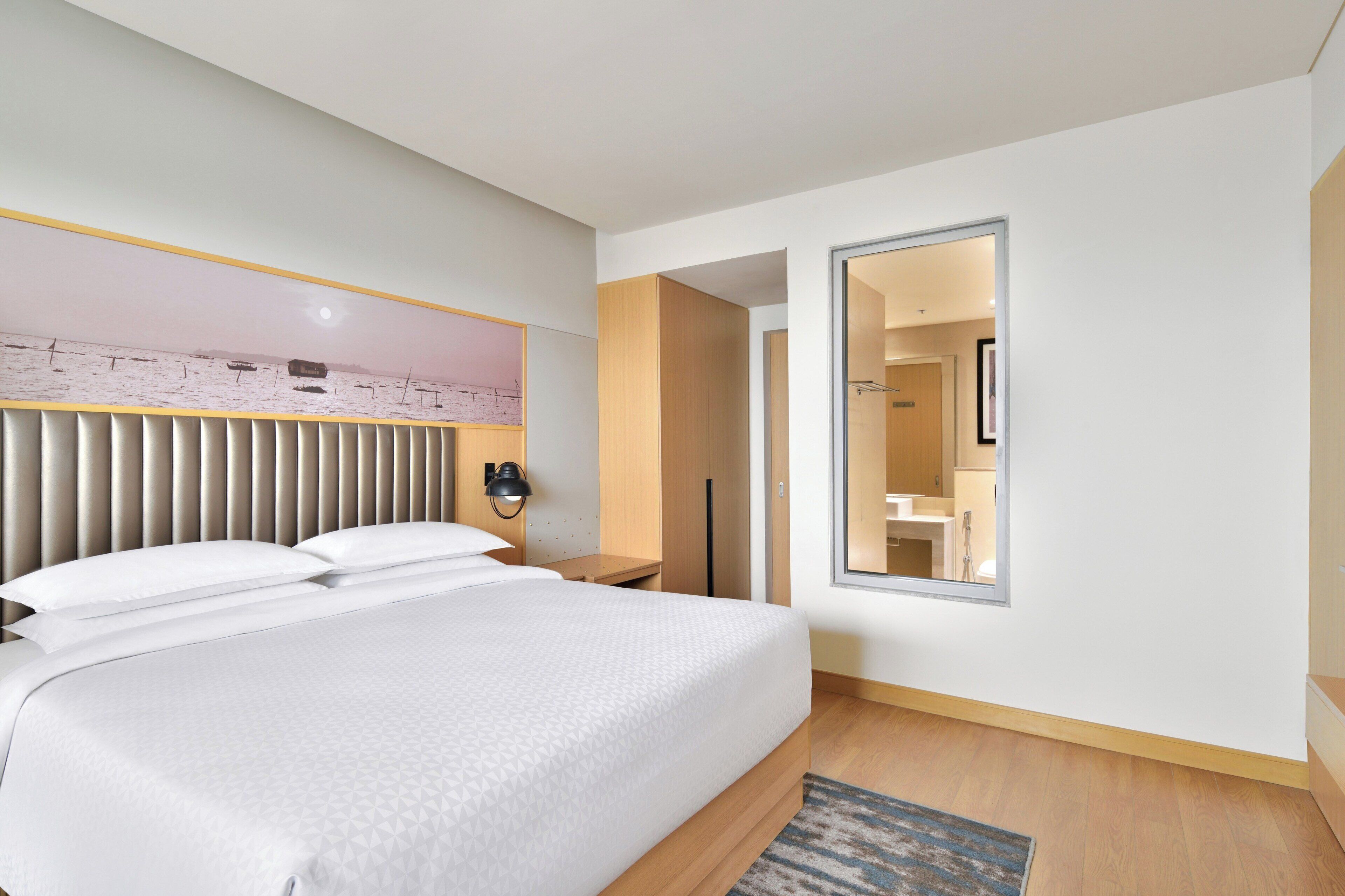 A modern hotel room with a large bed, a bedside table with a lamp, a mirror, and a view of the bathroom.