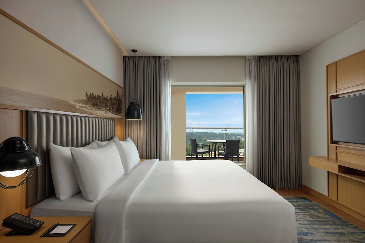 A hotel room with a bed, a desk with a lamp, a television, and a balcony with a table and chairs.