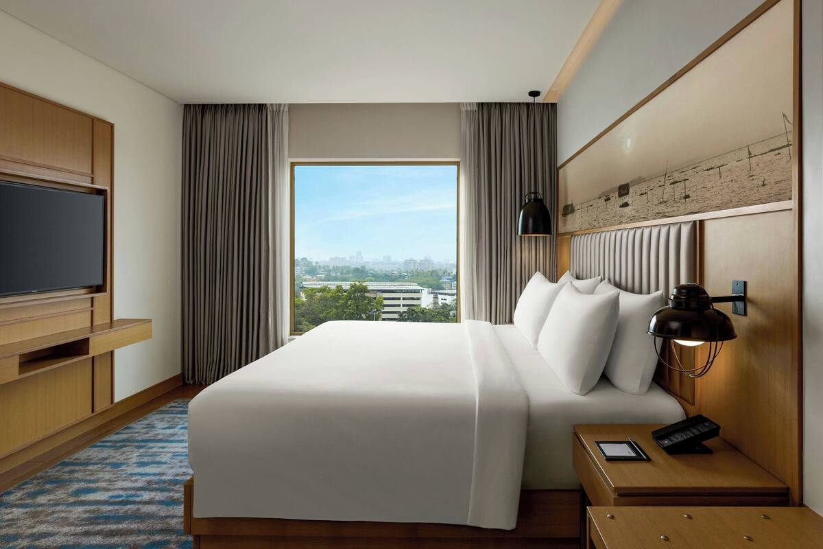 A hotel room with a large bed, a TV, a bedside lamp, and a view of the city.