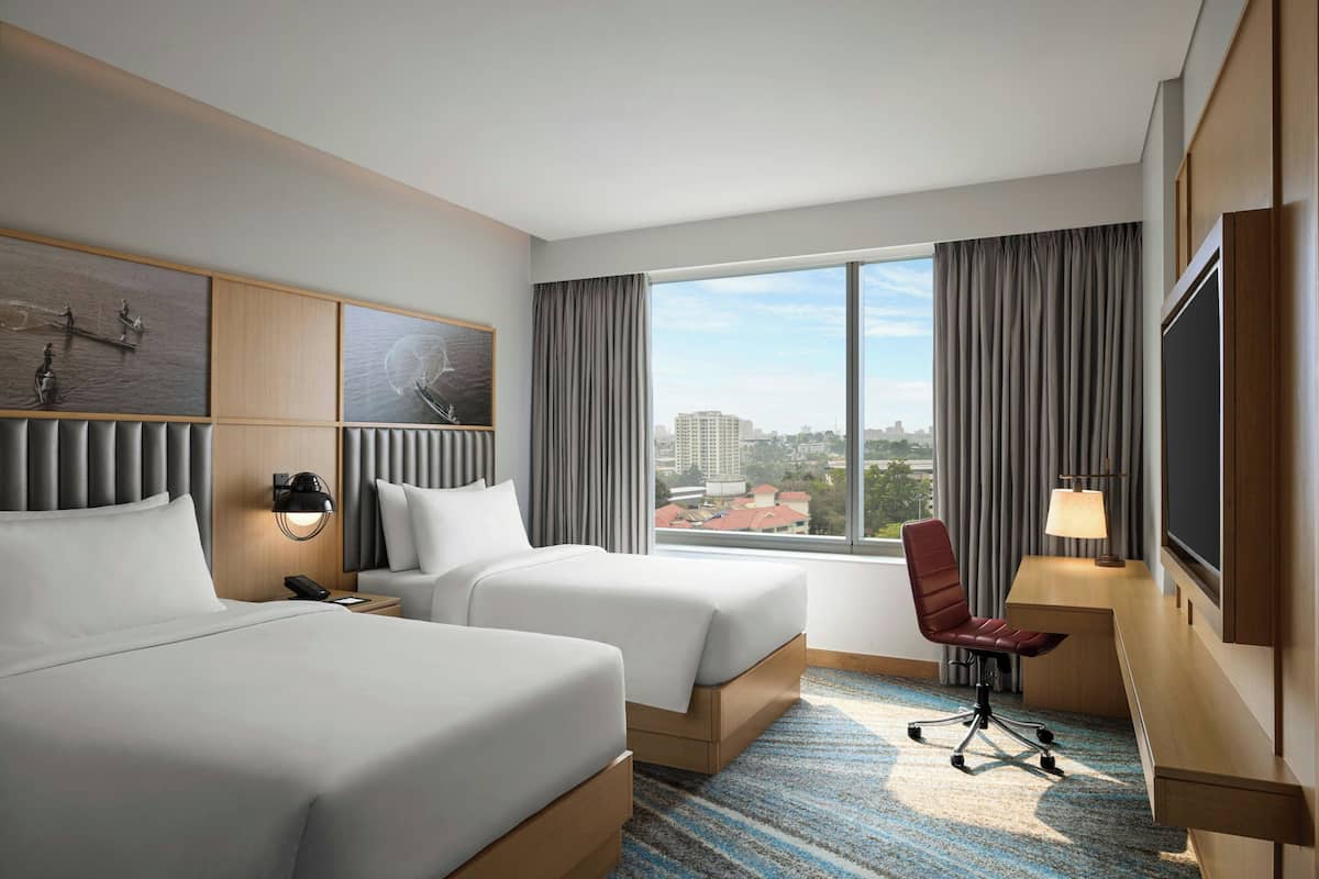 Une chambre d’hôtel avec deux lits, un bureau, une chaise, une télévision et une grande fenêtre donnant sur la ville.