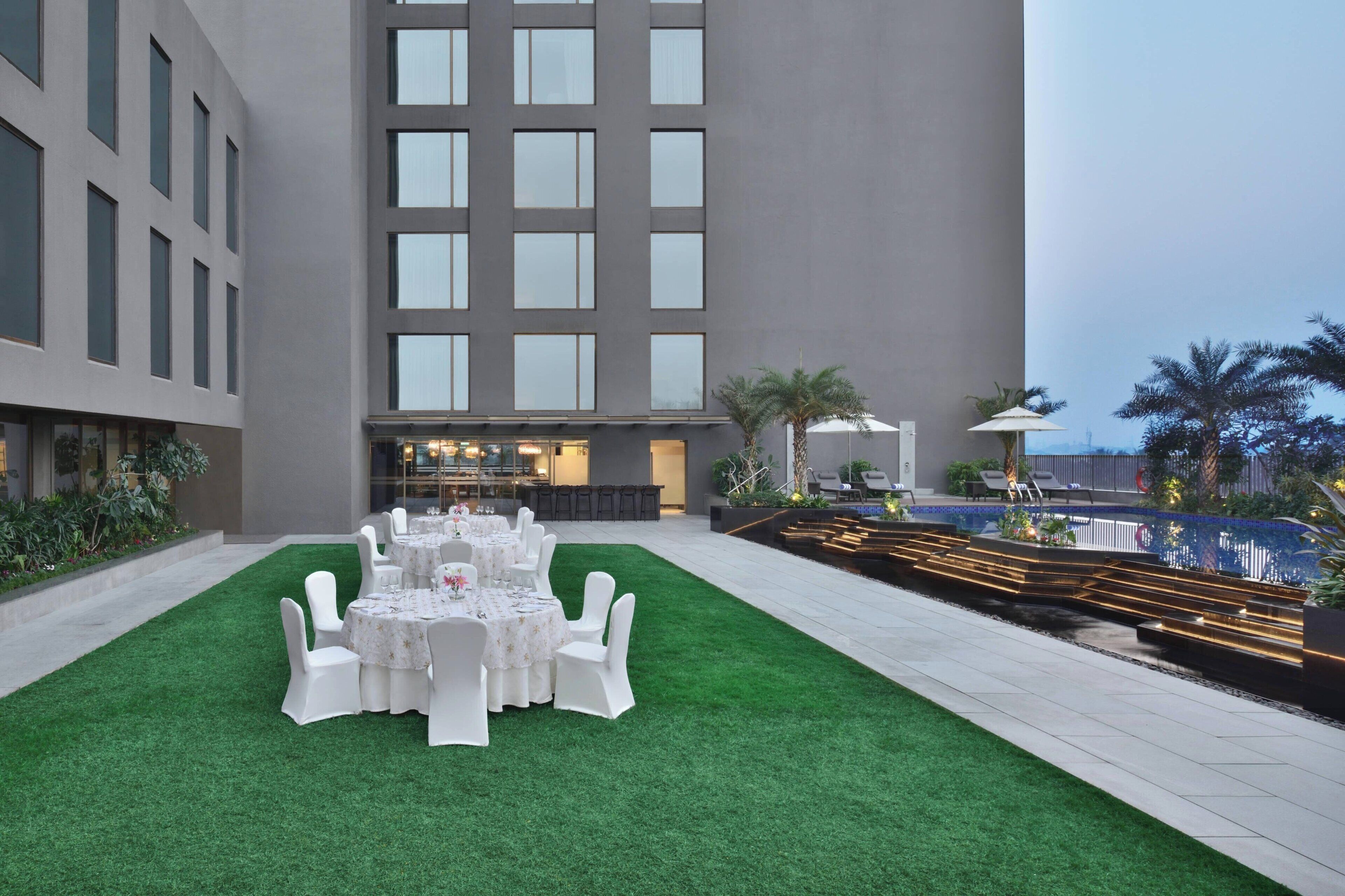 Un patio con zona de comedor dispuesto para un evento, rodeado por un edificio con amplias ventanas y una piscina.