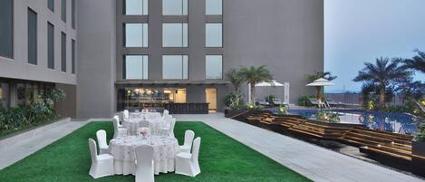 Un patio con zona de comedor dispuesto para un evento, rodeado por un edificio con amplias ventanas y una piscina.