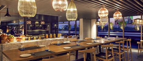 Un restaurante moderno con una larga mesa de madera, lámparas colgantes y vistas de la ciudad a través de amplios ventanales.