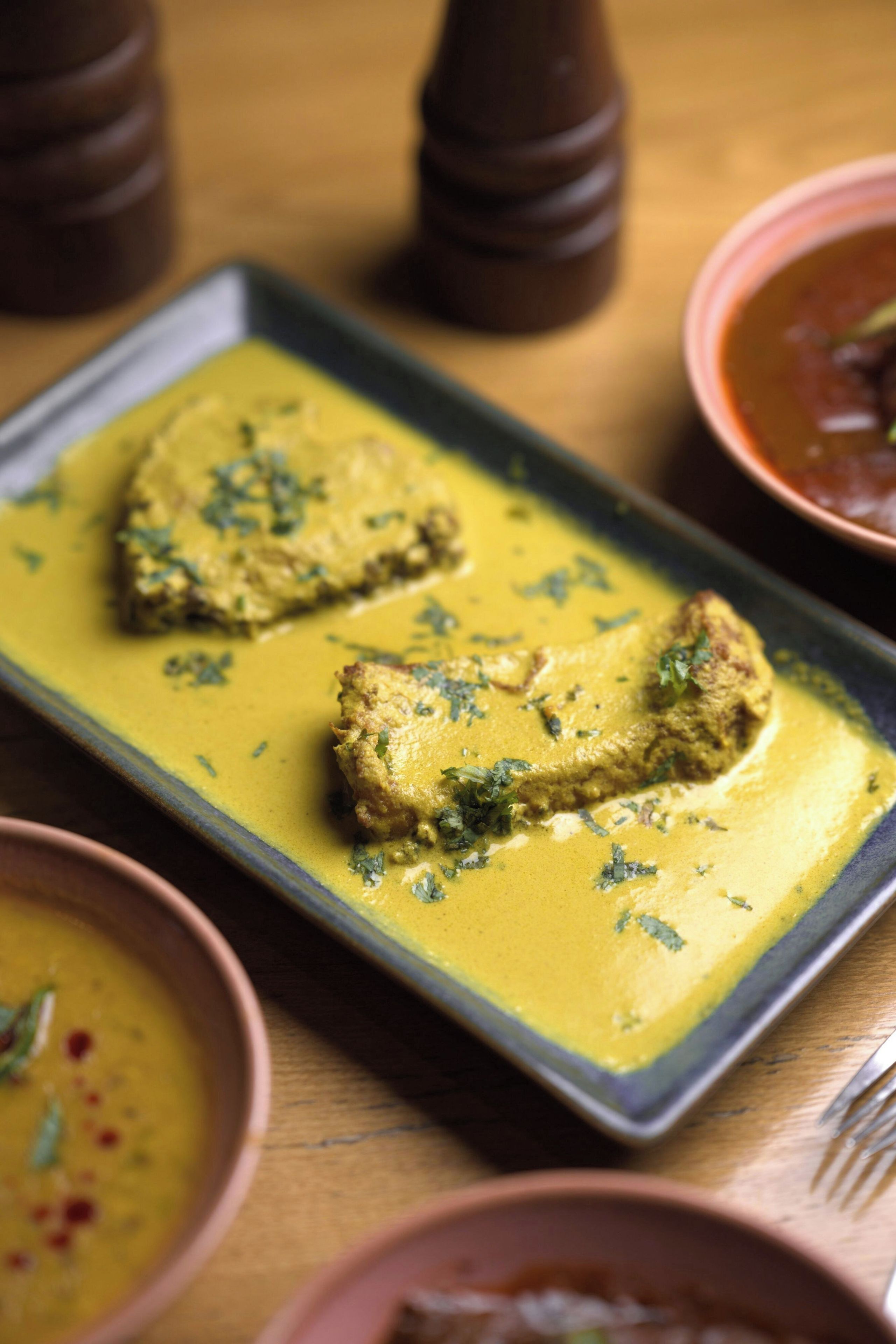 A rectangular plate with two pieces of food covered in a yellow sauce, garnished with green herbs, on a wooden table.
