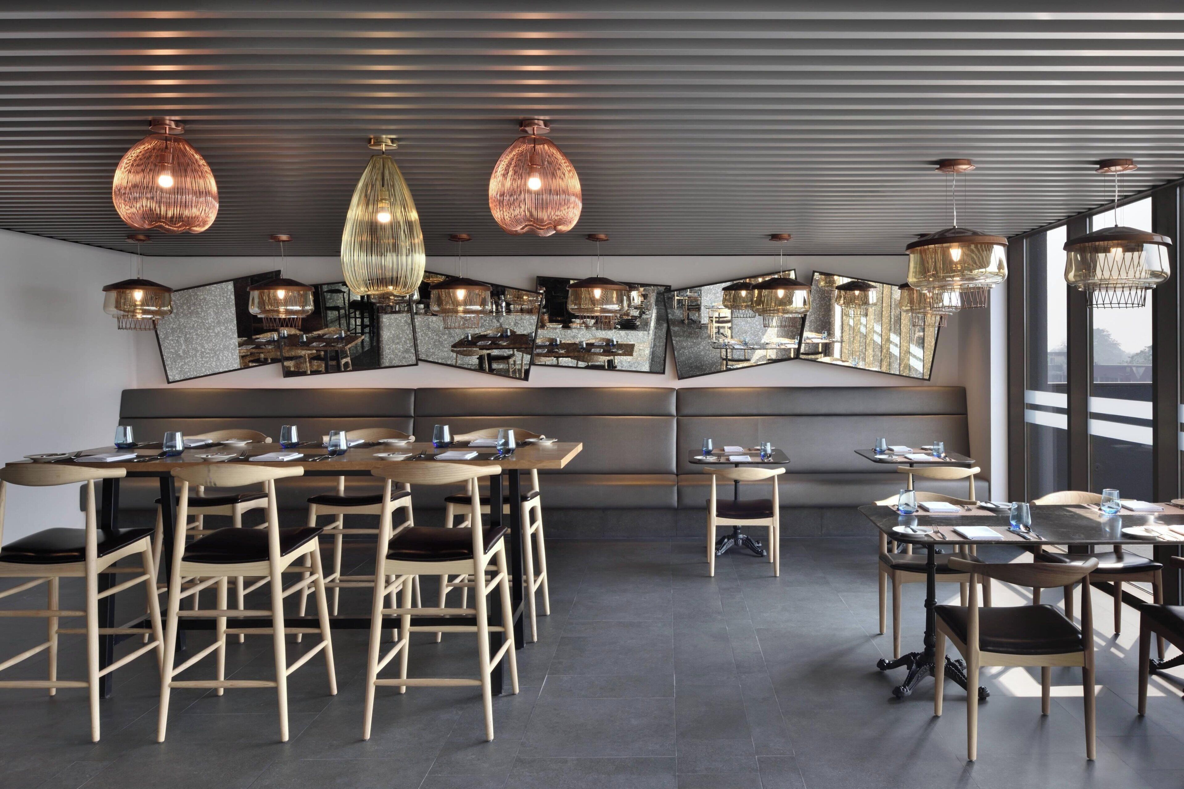 A modern restaurant with a central seating area, multiple hanging pendant lights, and large windows providing natural light.