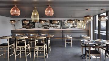 A modern restaurant with a central seating area, multiple hanging pendant lights, and large windows providing natural light.