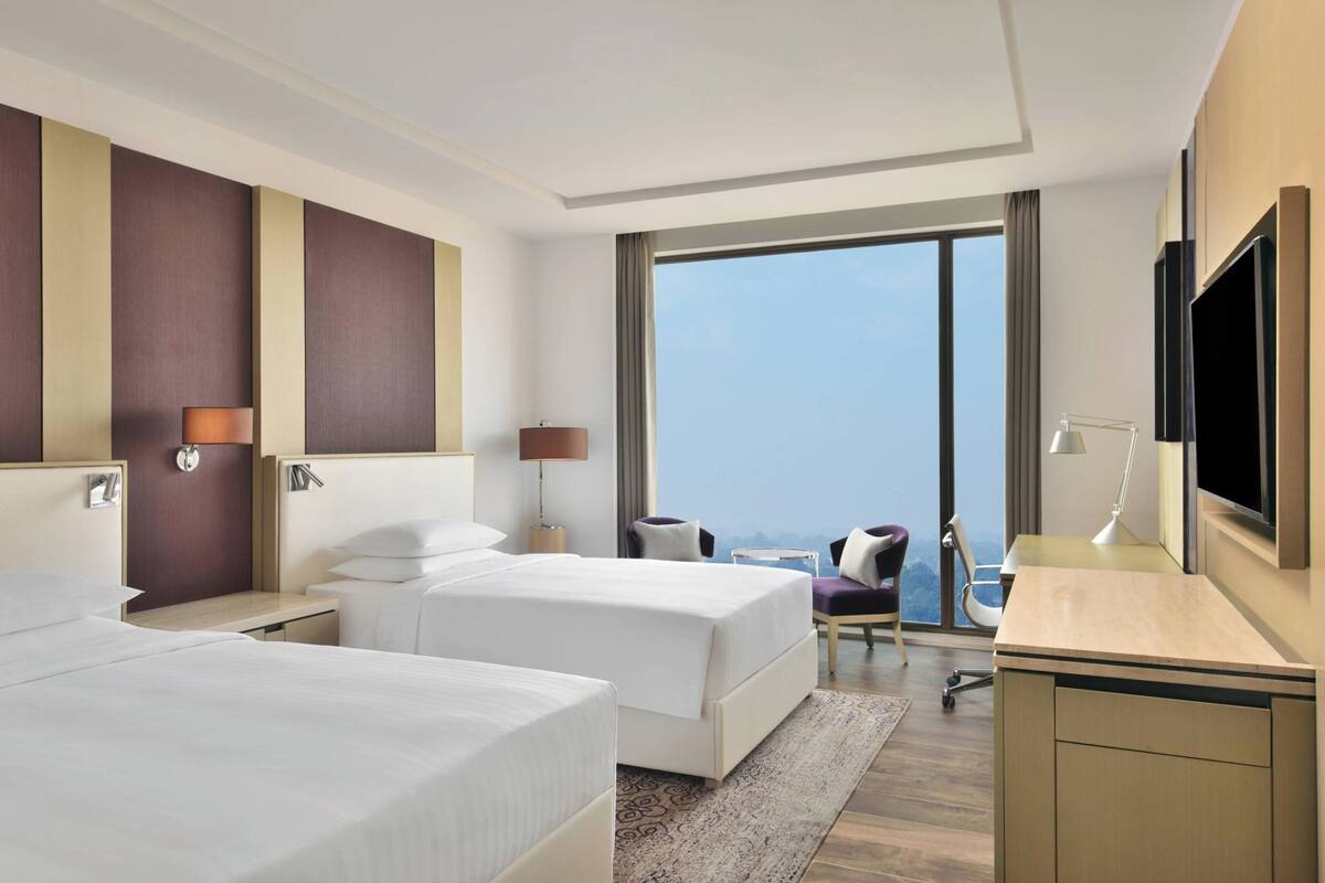 A hotel room with two beds, a desk, a TV, and a large window offering a city view.