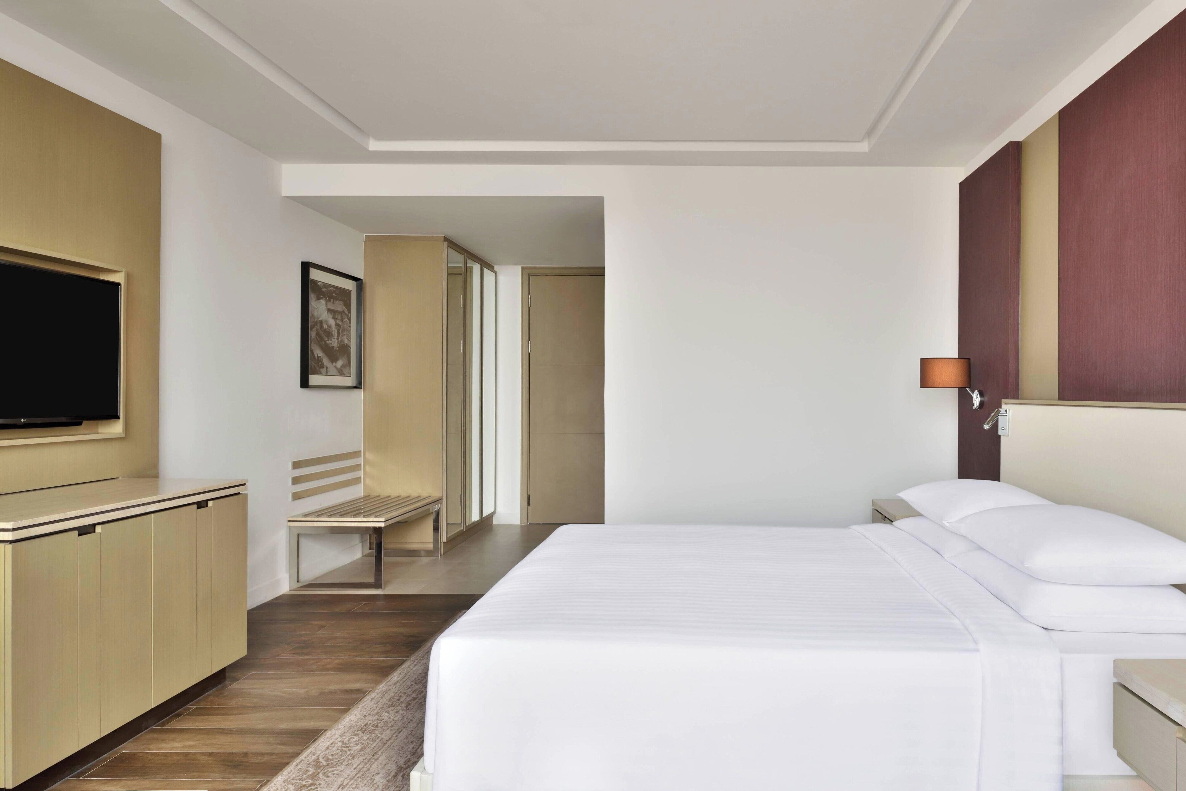 A hotel room with a bed, a television, a bench, and a wall-mounted lamp.