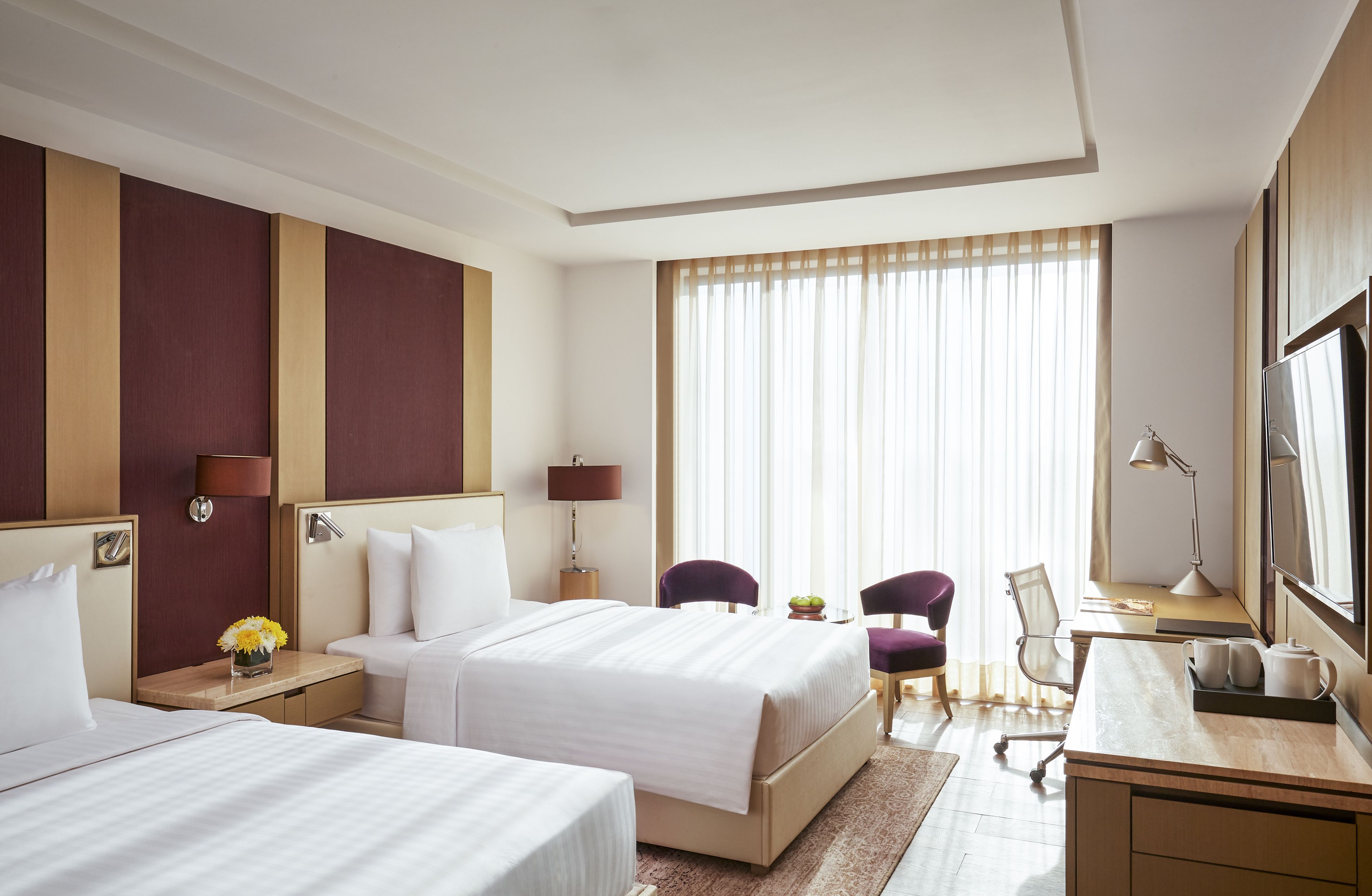 A hotel room with two beds, a desk, a chair, a TV, and a large window with curtains.
