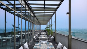 Un restaurante en la azotea con una larga mesa dispuesta para comer, rodeado de paredes de cristal y vistas a la ciudad.