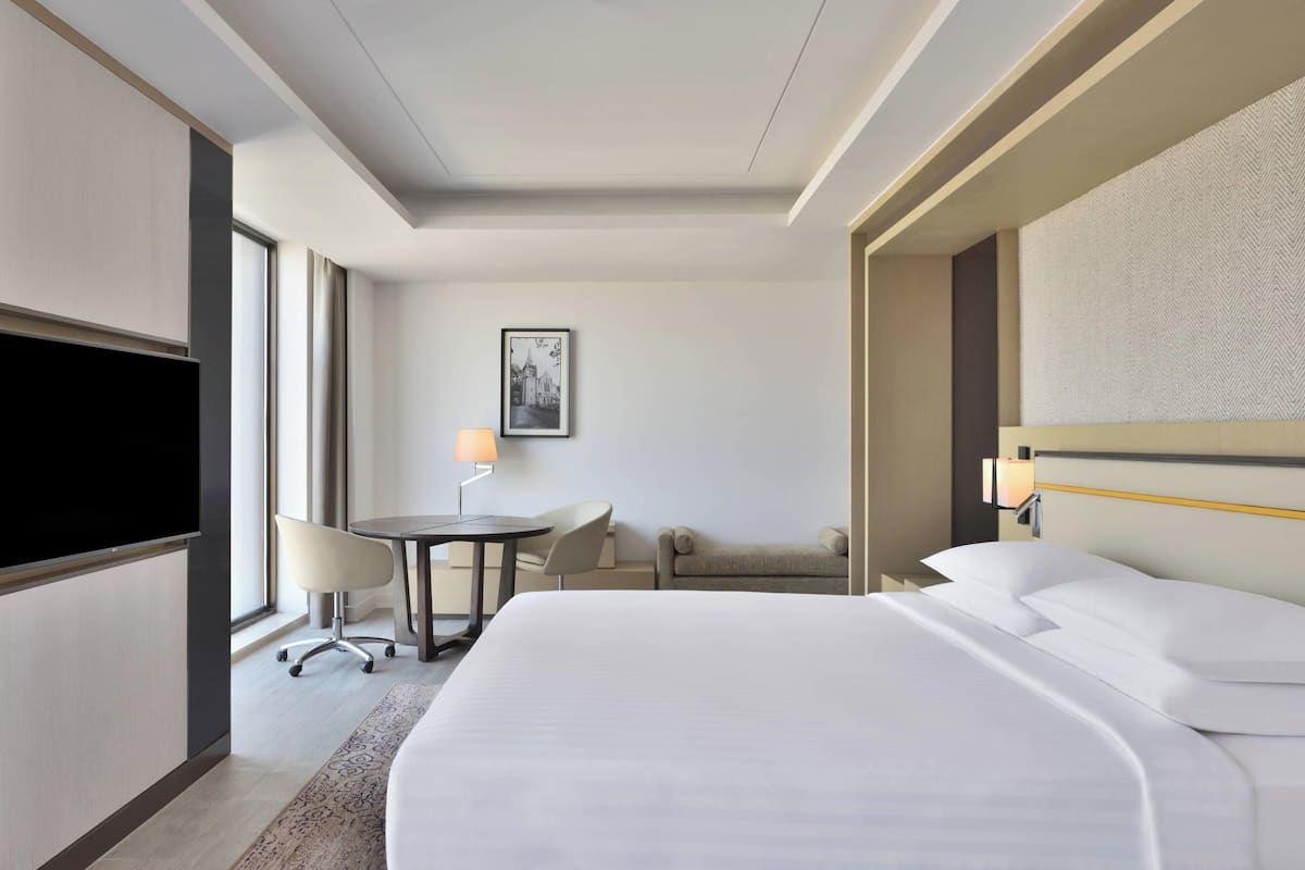A hotel room with a large bed, a flat-screen TV, a desk with a chair, a small table with a lamp, and a sofa.
