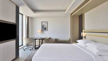 A hotel room with a large bed, a flat-screen TV, a desk with a chair, a small table with a lamp, and a sofa.