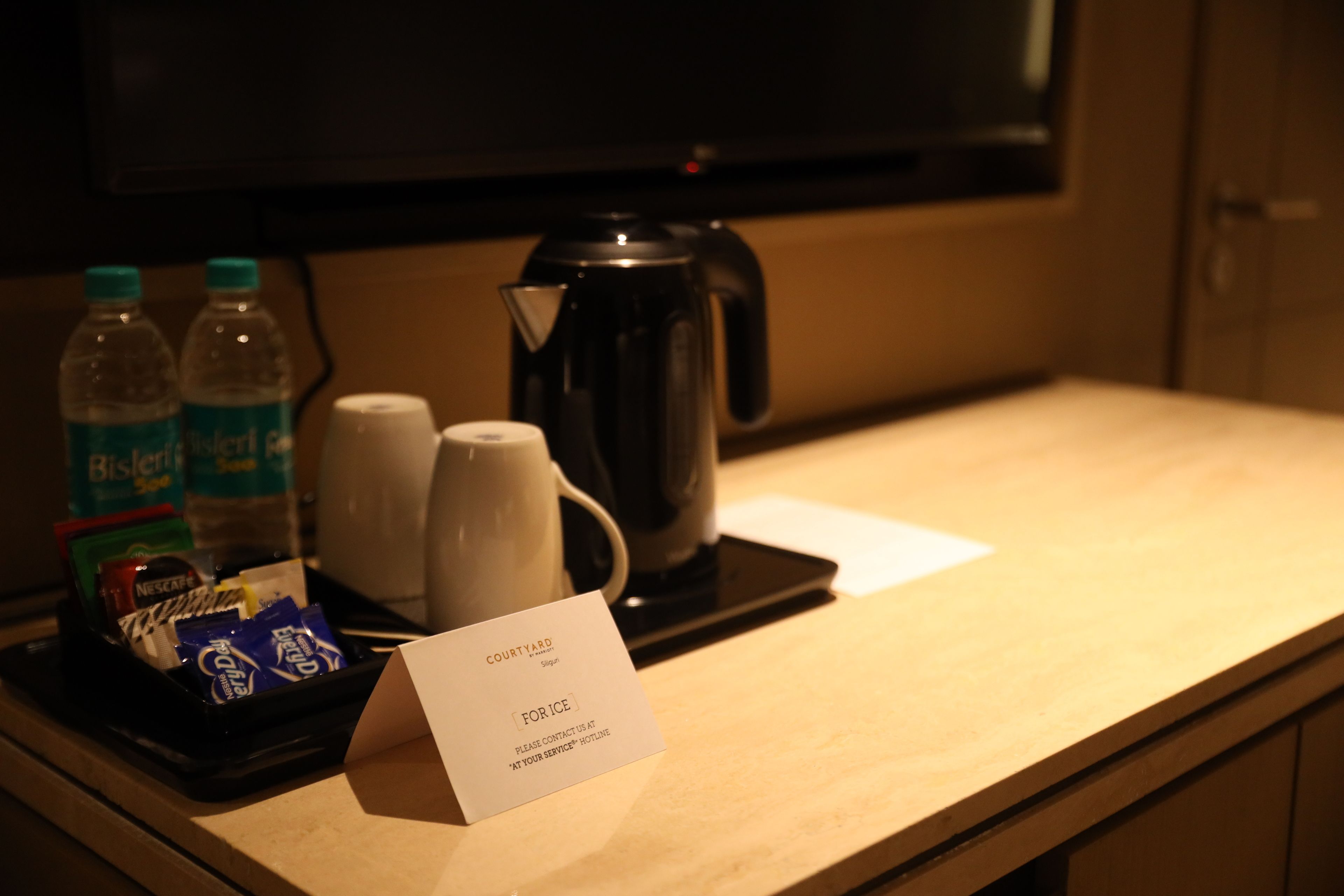 Bancone della reception di una camera d'albergo con una macchina per il caffè, due bottiglie d'acqua, una tazza da caffè e un vassoio con snack. Un cartello riporta le scritte 'COURT YARD' e 'FORICE'.