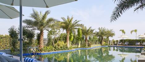 A poolside area with lounge chairs, umbrellas, and a clear pool reflecting palm trees and a fence.