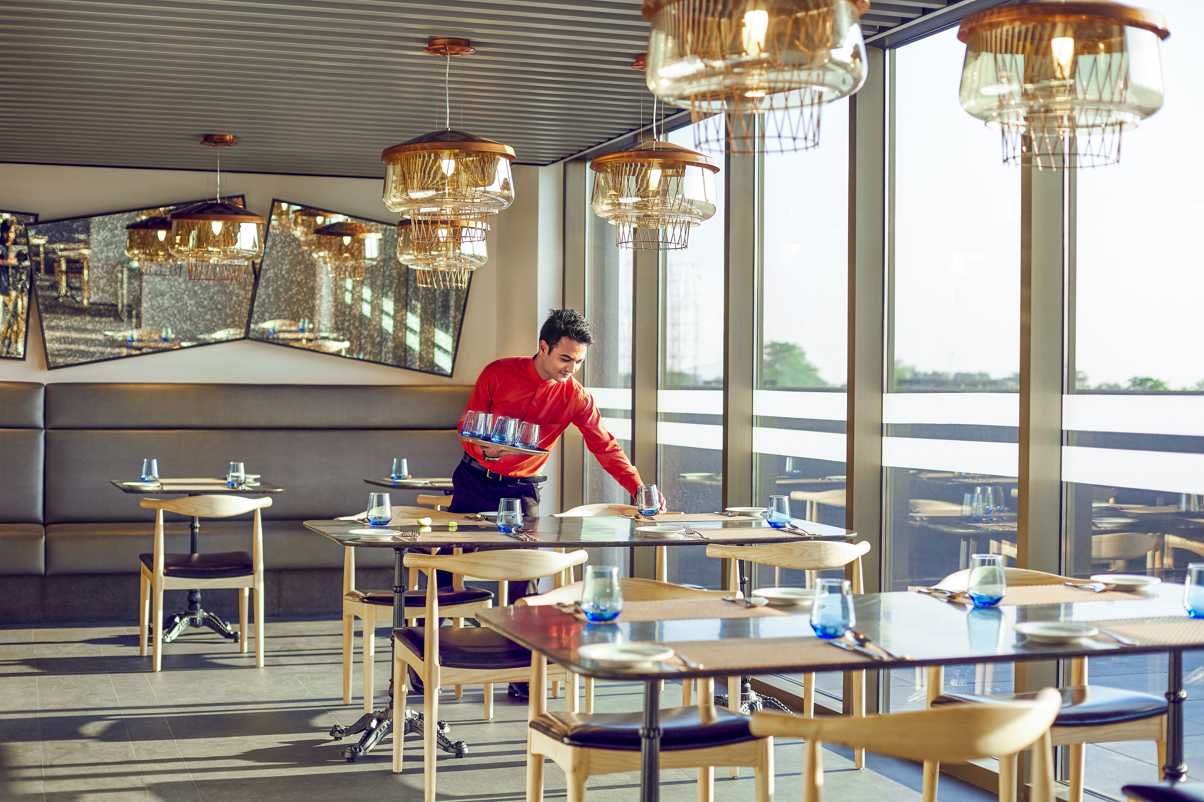 Un restaurante moderno con mesas y sillas de madera, ventanas amplias y lámparas colgantes.