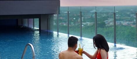 Un couple est assis au bord d’une piscine à débordement, avec une vue sur la ville. On aperçoit un bâtiment moderne aux fenêtres en verre et un ciel dégagé.