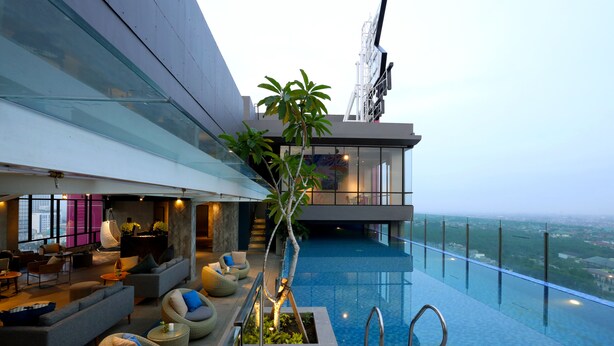 A modern infinity pool with a glass barrier, a lounge area with sofas and a bar, and a view of the cityscape. - The Zuri Pekanbaru (Pekanbaru)