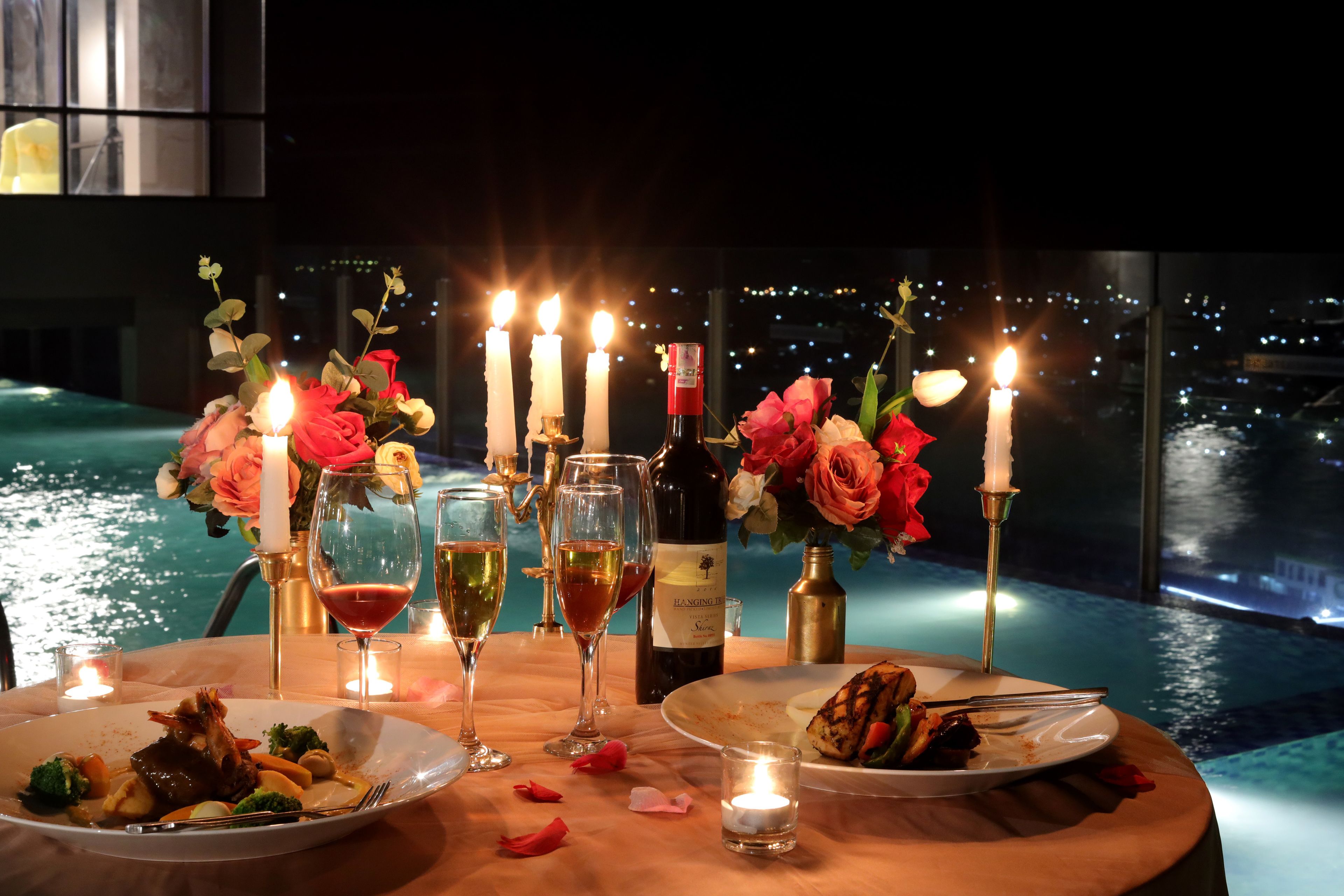 A romantic dinner setup by a pool with candles, flowers, and wine glasses.