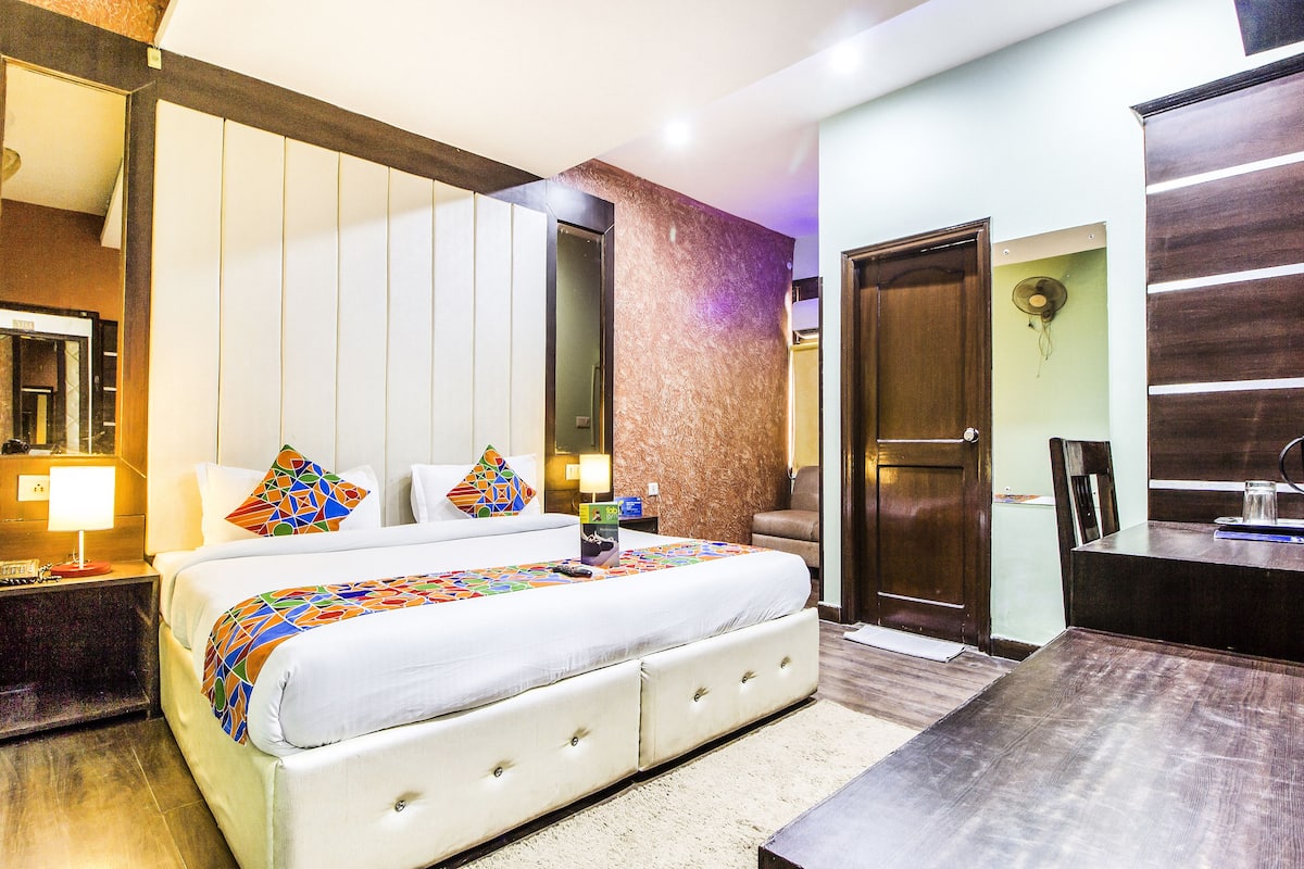 A hotel room with a large bed, a desk, and a chair. There is a door leading to a bathroom and a mirror on the wall.