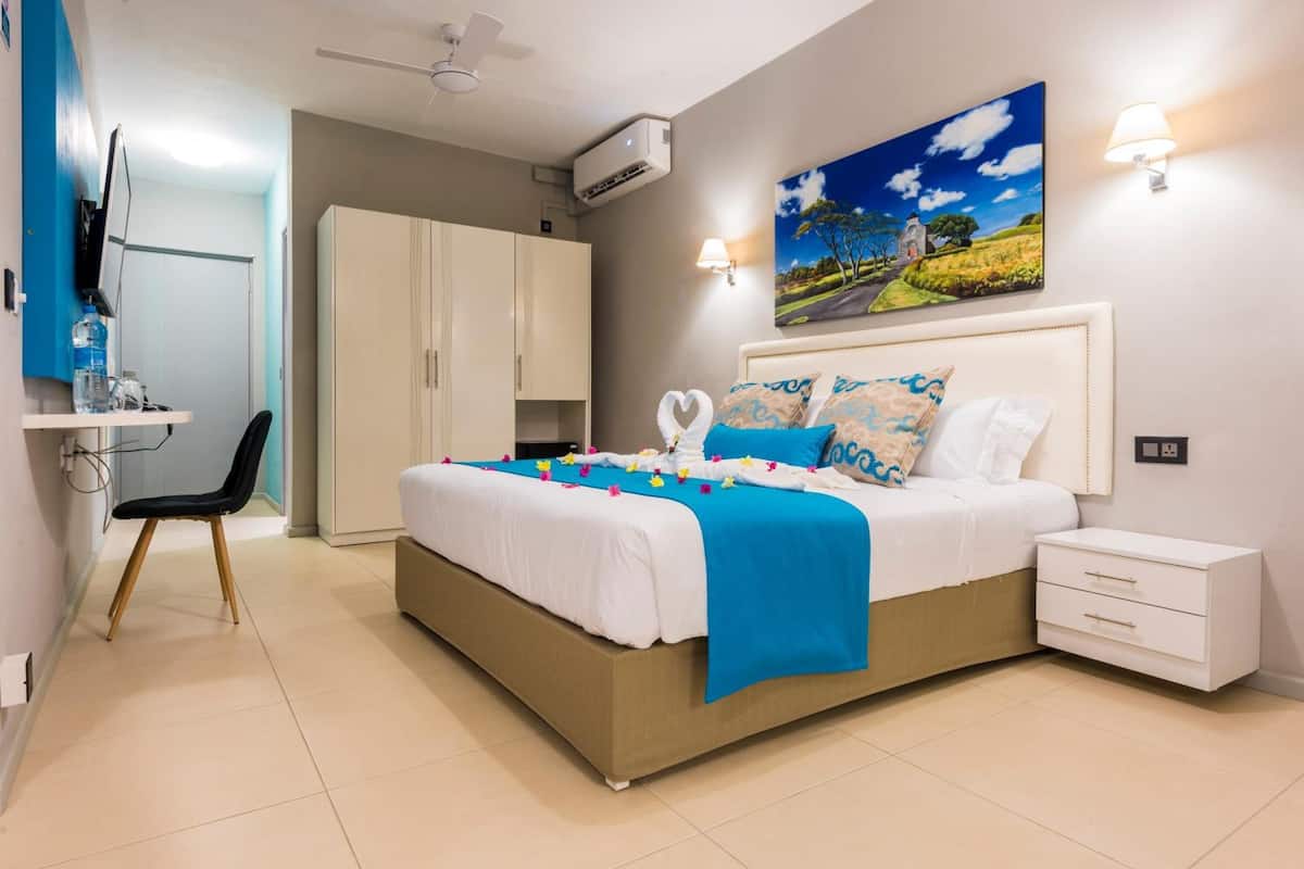 Um quarto com cama, mesas de cabeceira, escrivaninha com cadeira, ar condicionado e um quadro na parede.
