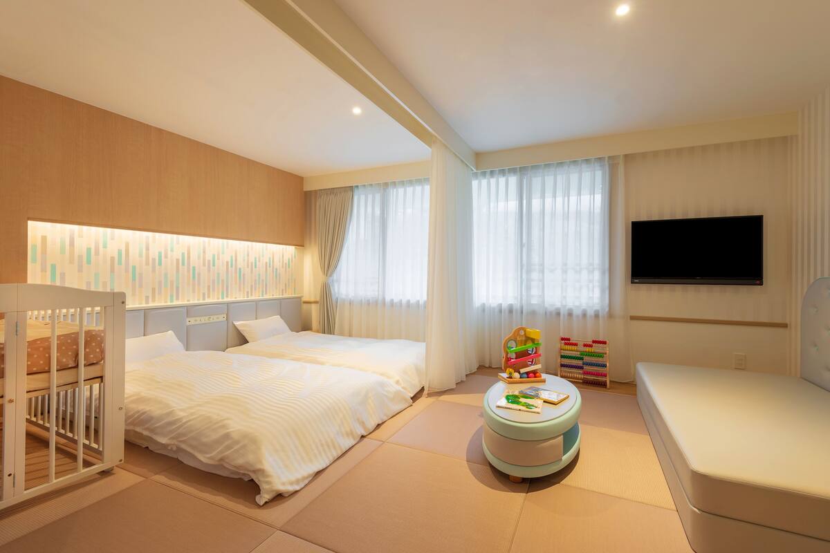 A hotel room with two beds, a TV, a crib, and a play area with toys.