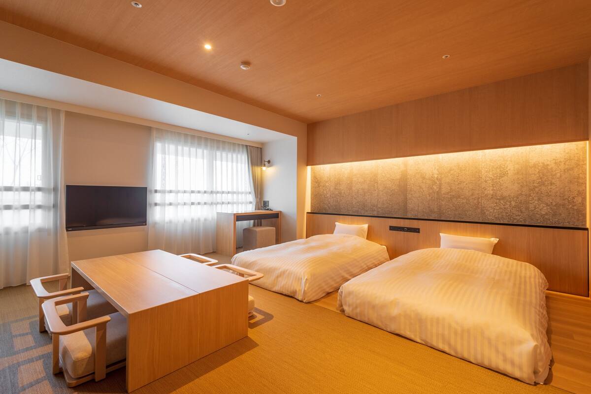 A modern hotel room with two beds, a wooden desk, and a large window with curtains.