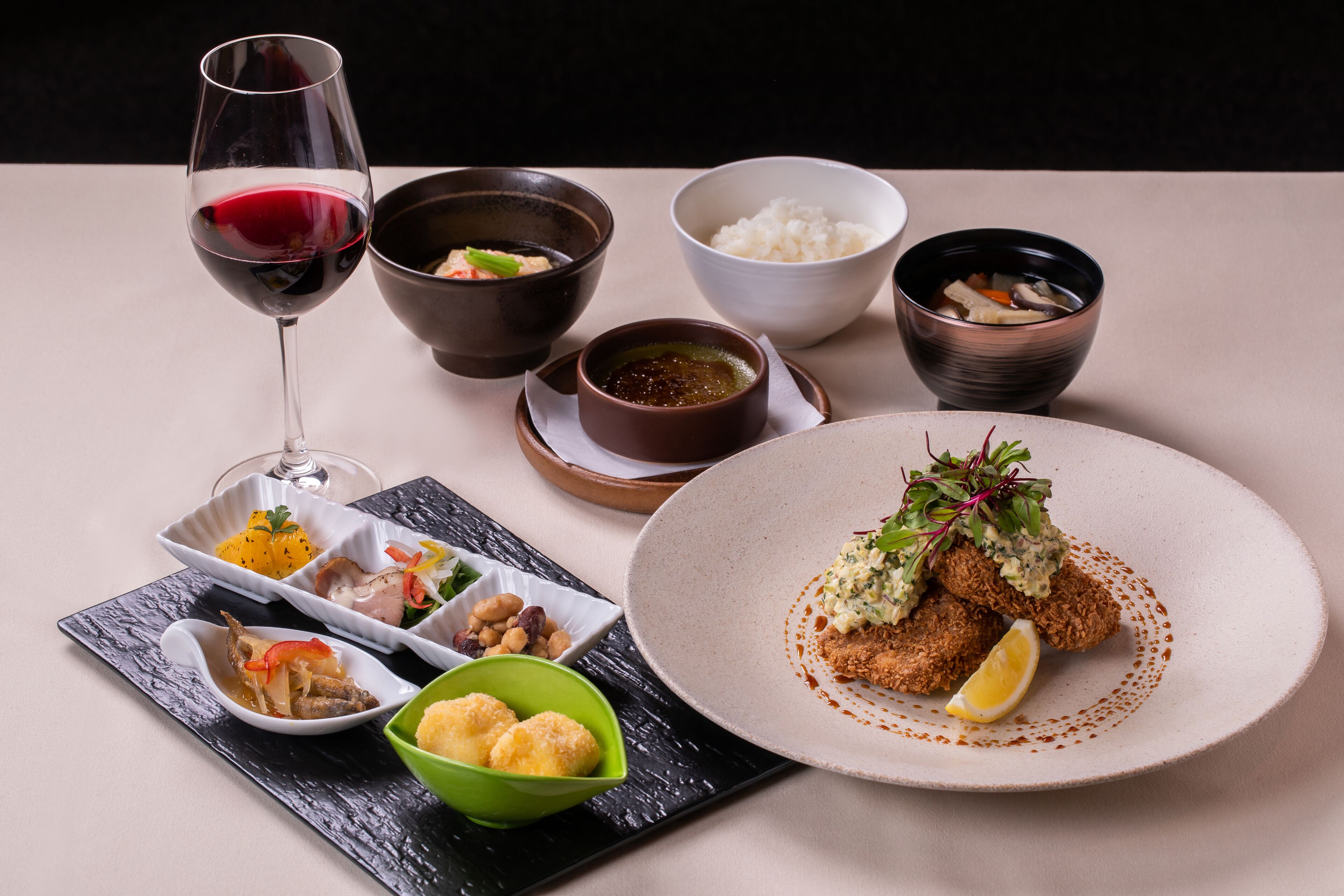 A table set with various dishes, including a glass of red wine, bowls of rice, soup, and a plate with a breaded item garnished with greens and a lemon wedge.