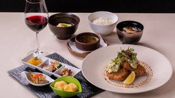 A table set with various dishes, including a glass of red wine, bowls of rice, soup, and a plate with a breaded item garnished with greens and a lemon wedge.