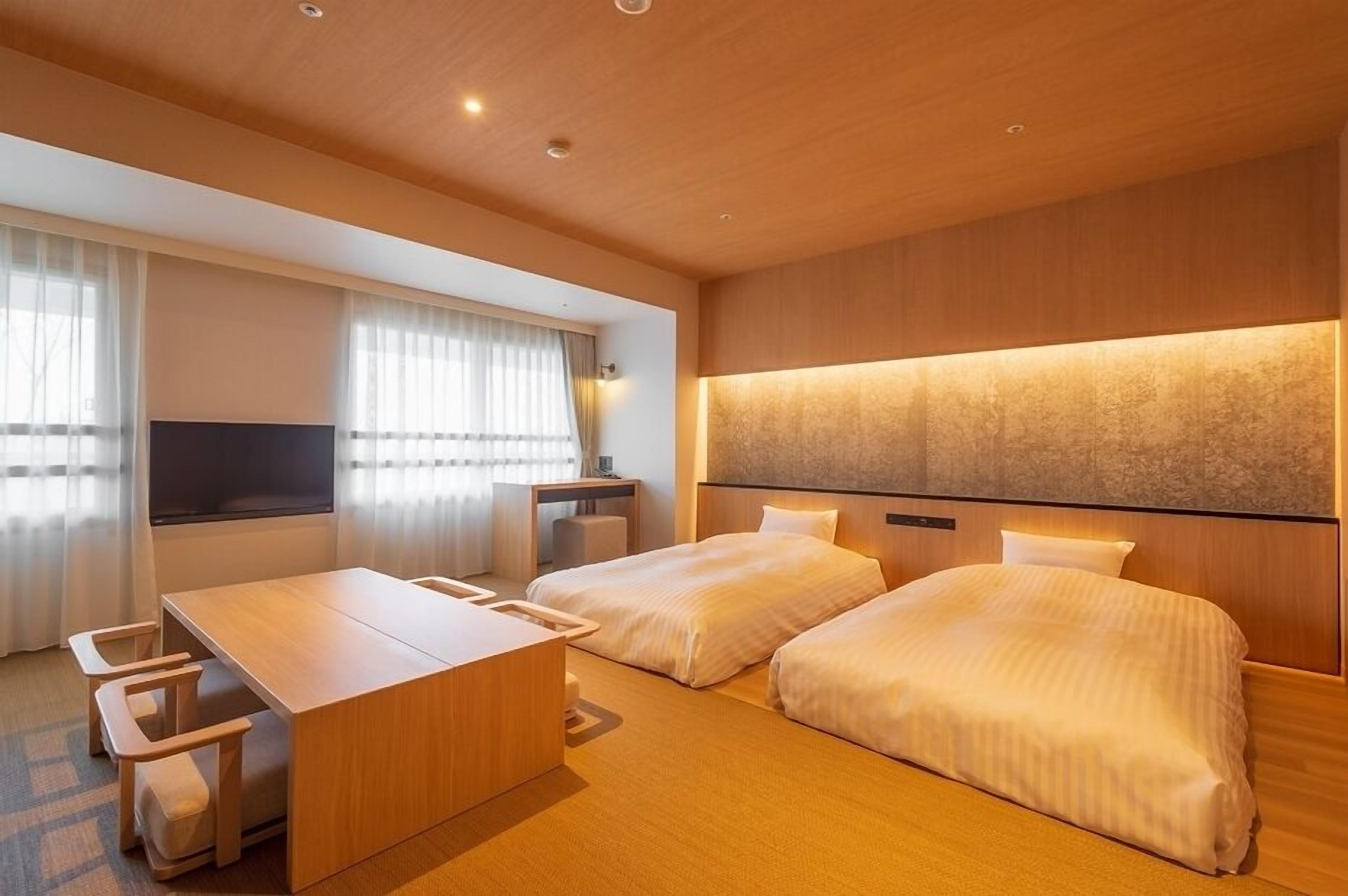 A hotel room with two beds, a wooden desk, and a large window with curtains.